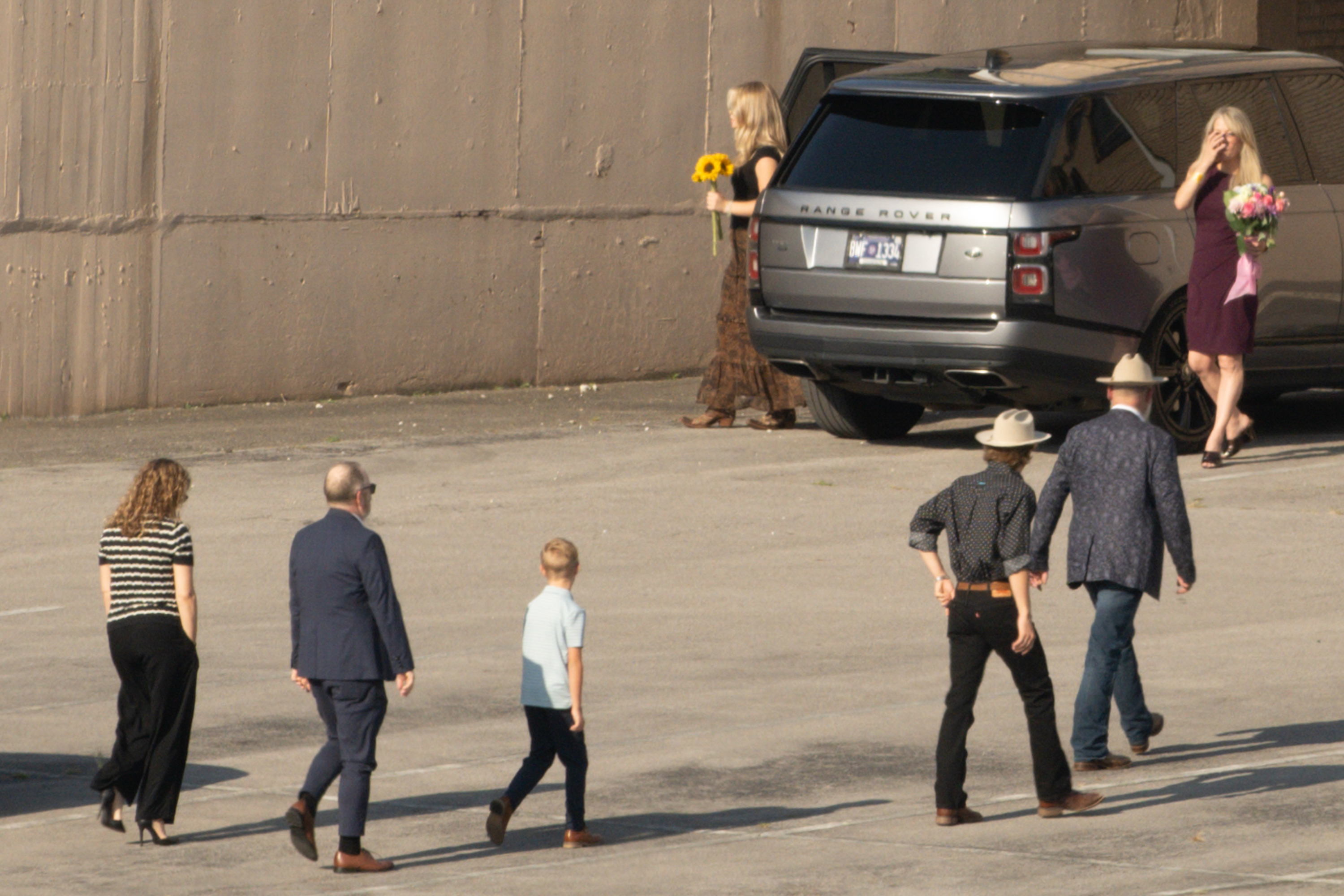Several people, two carrying flowers, walking across a parking lot away from a Range Rover.