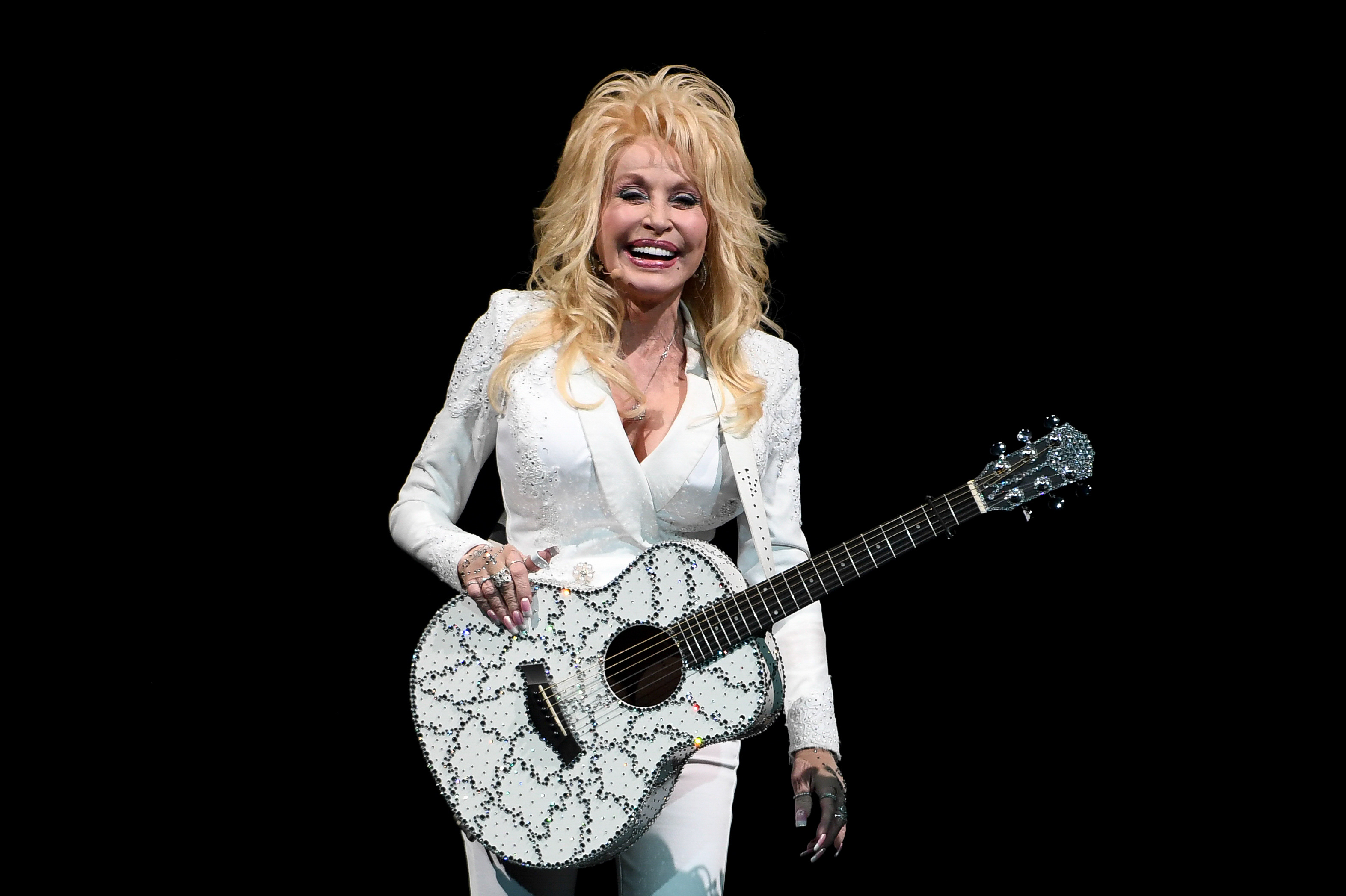 Dolly Parton in a white suit, holding a sparkling white guitar, smiling on stage.