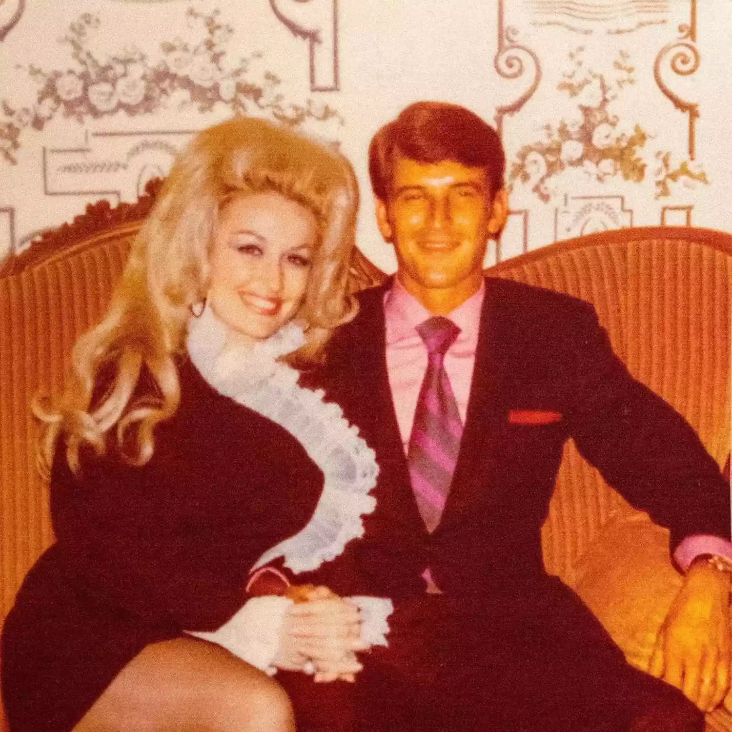 Dolly Parton and her husband Carl Thomas Dean sitting together, with Parton smiling brightly at the camera.