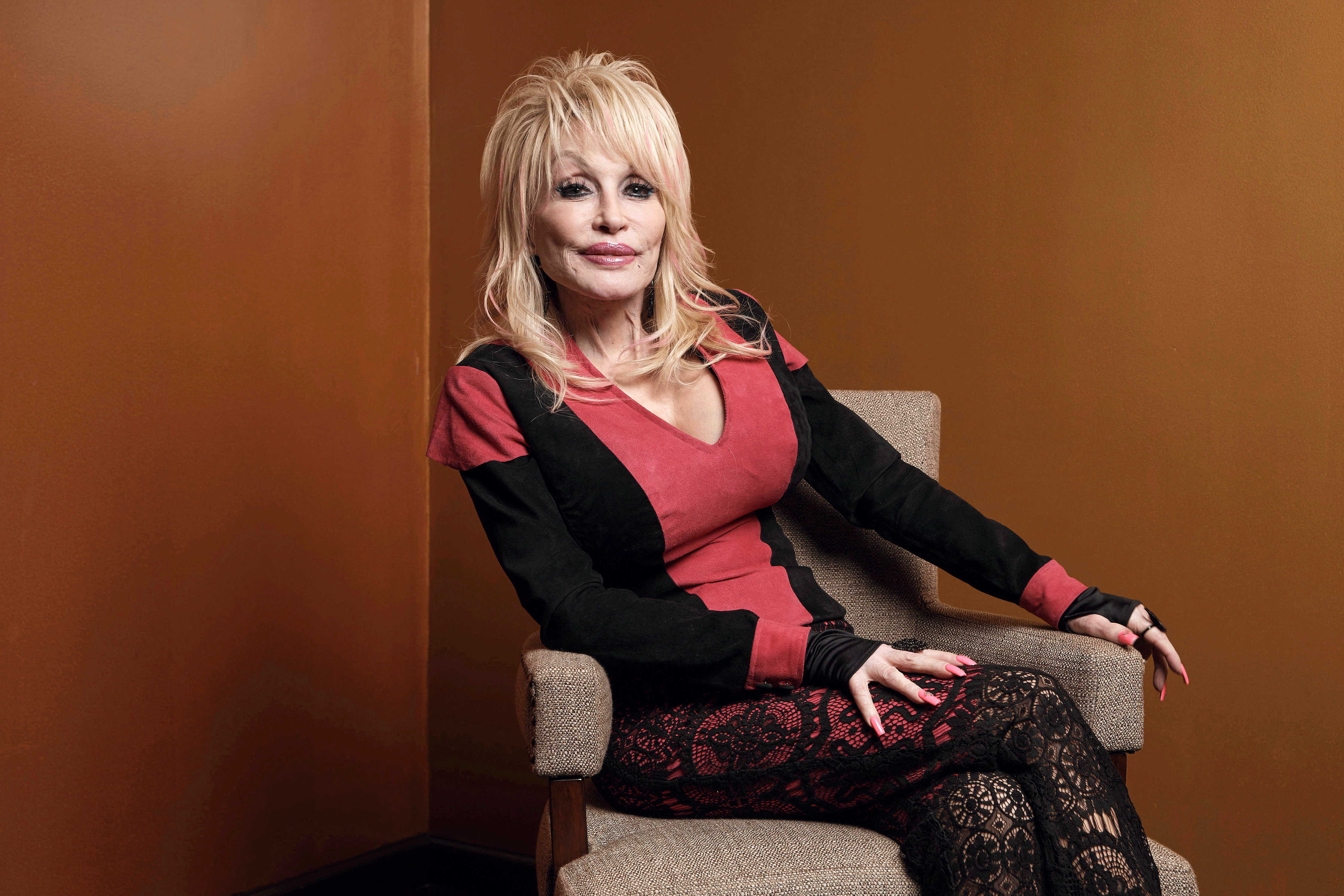 Dolly Parton in a red and black outfit, sitting in a chair.